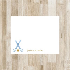 May include: A rectangular white stationery card featuring a graphic of two crossed light blue field hockey sticks with a gold ball at the base. The name Jessica Cassidy is printed in gold serif lettering on the bottom right side of the card.