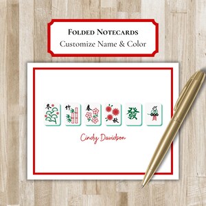 May include: White folded notecard with a red border, featuring Mahjong tile designs and the name "Cindy Davidson" in red. A gold pen rests on the card. The text "Folded Notecards Customize Name & Color" is at the top.