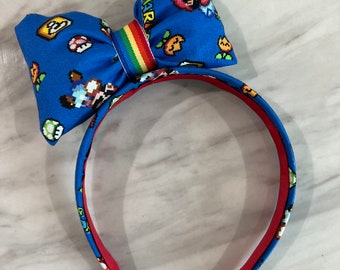 May include: A blue headband with a large bow featuring a pixelated Mario character design. The bow has a rainbow stripe in the center and the text "MARIO" is visible on the bow.