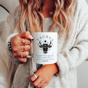 May include: White ceramic mug featuring the word "TAURUS" in blue script above a black bull illustration with celestial accents. The mug also includes zodiac traits and planet information. A perfect gift for astrology enthusiasts.