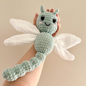May include: A handmade crochet dragonfly toy in soft pastel green and white. The dragonfly has large white wings, a rounded body, and a smiling face with black eyes and pink cheeks. The toy is made of a plush, textured yarn.
