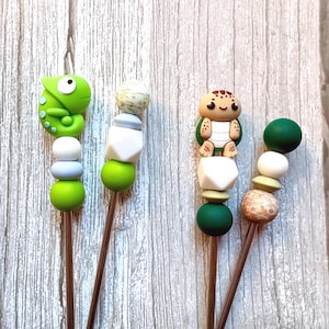 May include: Two decorative skewers with colourful beads and animal toppers. One skewer features a green chameleon, the other a turtle. The beads are white, green, and blue, with a wooden stick.