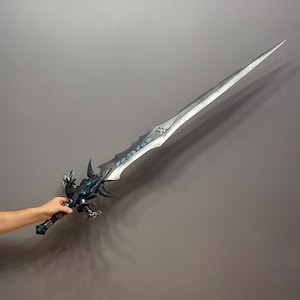Pu&ograve; includere: Una grande spada fantasy argentata con intricati dettagli neri e blu. La lama &egrave; lunga e appuntita, con un'elsa decorativa. La spada &egrave; tenuta su uno sfondo grigio.