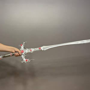May include: A silver sword with a decorative handle and red gems. The sword is held by a person's hand.