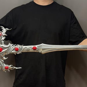 May include: A silver sword with red gems, held by a person wearing a black shirt. The sword has a detailed design with a dragon-like handle.