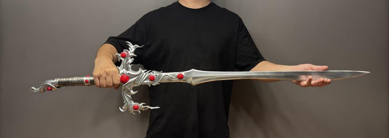 May include: A silver sword with red gems, held by a person wearing a black shirt. The sword has a detailed design with a dragon-like handle.