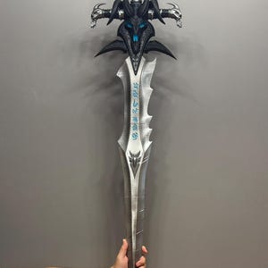 Pu&ograve; includere: Una grande spada fantasy con una lama argentata e un'elsa nera e blu dettagliata. L'elsa presenta un design a teschio e dettagli ornati. La spada &egrave; tenuta da una persona.