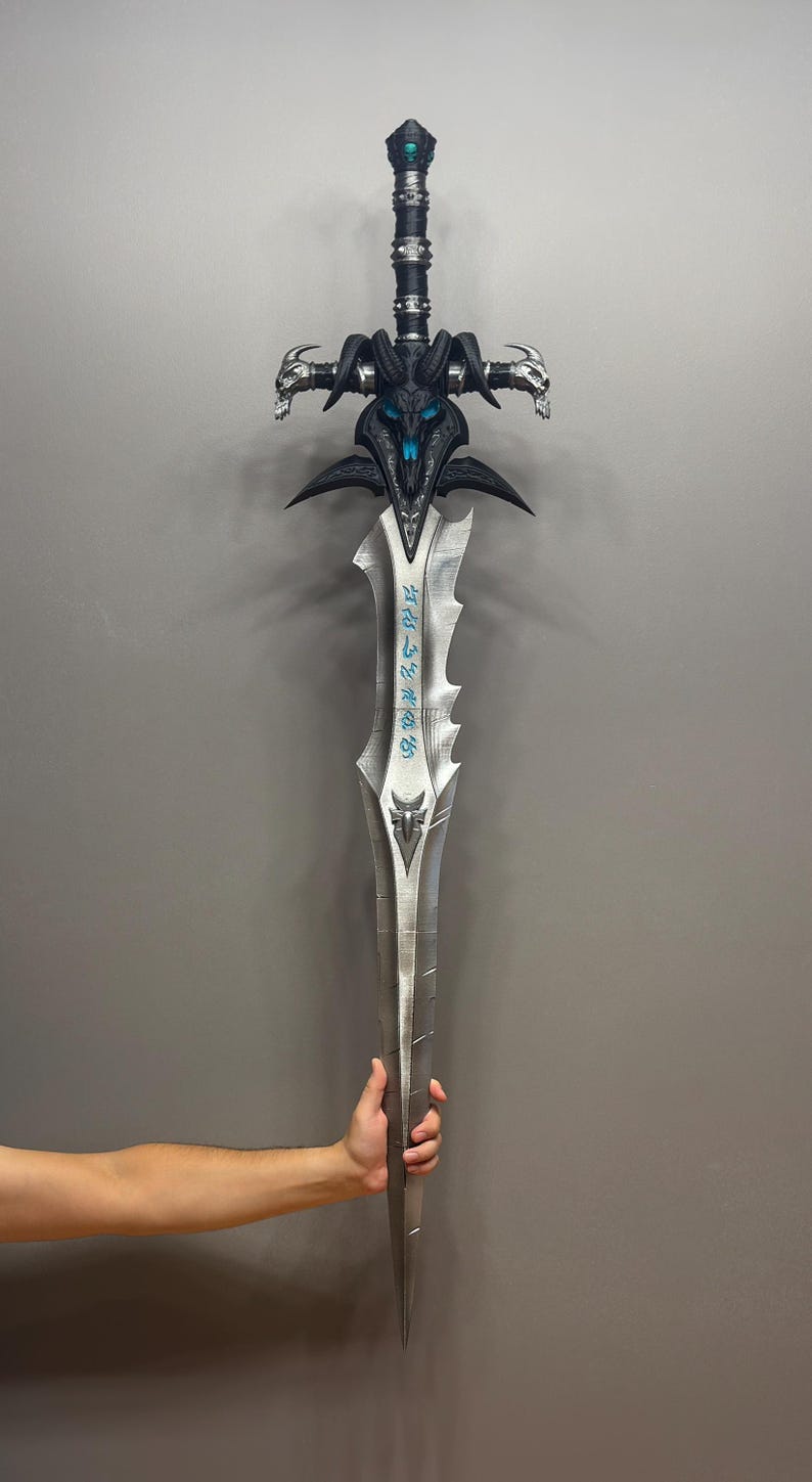 Pu&ograve; includere: Una grande spada fantasy con una lama argentata e un'elsa nera e blu dettagliata. L'elsa presenta un design a teschio e dettagli ornati. La spada &egrave; tenuta da una persona.
