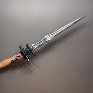 Pu&ograve; includere: Una spada fantasy dettagliata con una lama argentata e un'elsa nera e blu intricata. La lama presenta un design decorativo con dettagli blu. L'elsa presenta un design ornato. La spada &egrave; tenuta su uno sfondo grigio.