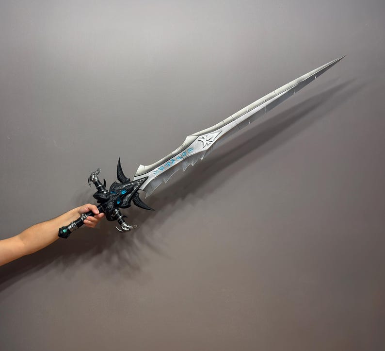Pu&ograve; includere: Una spada fantasy dettagliata con una lama argentata e un'elsa nera e blu intricata. La lama presenta un design decorativo con dettagli blu. L'elsa presenta un design ornato. La spada &egrave; tenuta su uno sfondo grigio.