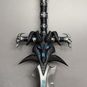 Pu&ograve; includere: Spada fantasy dettagliata con impugnatura e guardia nere, con disegni di teschi e corna di ariete. La lama &egrave; argentata con testo blu e l'elsa ha un pomo a forma di teschio. Il design generale &egrave; intricato e gotico.