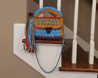 May include: A colorful, handmade handbag with a braided design. The bag features a variety of colors, including blue, orange, yellow, and gray. It has a shoulder strap and a decorative tassel. The bag is sitting on a wooden surface.