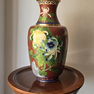 May include: A decorative vase with a maroon background and floral designs in yellow, blue, and white. The vase has a gold rim and a detailed pattern. The vase is on a wooden stand.