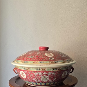 May include: A red and white ceramic serving bowl with a lid. The bowl features a floral pattern and Chinese characters. The lid has a red knob. The bowl is sitting on a wooden stand.