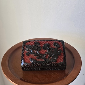 Può includere: Una scatola nera rettangolare con interno rosso intrecciato e intricate sculture floreali. La scatola è su un vassoio rotondo di legno. Le sculture raffigurano fiori e fogliame.