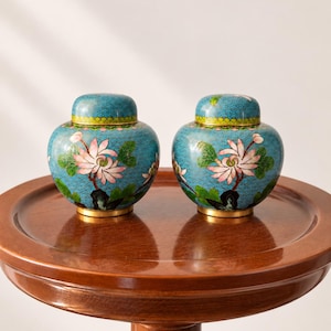Pode incluir: Um par de potes redondos com tampa a condizer, apresentando um fundo azul turquesa vibrante com um padrão de fio fino. Cada pote é decorado com flores de lótus cor-de-rosa e folhas verdes pintadas à mão, com rebordo e base dourados.