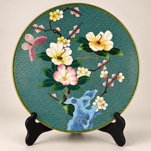 Pode incluir: Um prato decorativo redondo com fundo verde-azulado e um padrão de escamas finas. O design inclui uma borboleta rosa, flores brancas e amarelas, folhas verdes e uma formação rochosa azul. O prato tem uma borda fina de metal dourado.