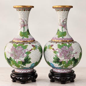 May include: A pair of cloisonné vases featuring a white background with a repeating cloud pattern, decorated with purple chrysanthemum flowers, green leaves, and small butterflies. Each vase has a gold-toned rim and sits on a black wooden stand.