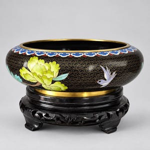 May include: A round cloisonné bowl with a black background featuring a gold cloud pattern, a large yellow peony, and a light blue bird. The bowl has a gold rim and sits on a dark, carved wooden stand with four feet. The interior is black.