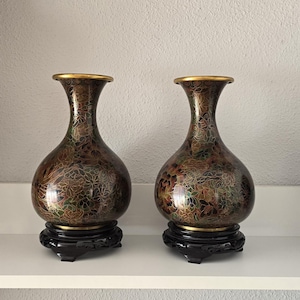May include: Two decorative vases with a floral pattern in shades of brown, green, and gold. The vases have a bulbous shape and a narrow neck, with a gold rim. They are displayed on black wooden stands.