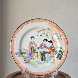 Op de afbeelding: Een decoratieve porseleinen bord met een handgeschilderde scène van drie vrouwen in traditionele kledij. Het bord heeft een gouden rand en een gedetailleerde rand. De vrouwen bevinden zich in een tuin met bomen en een kleine brug. Het bord wordt op een transparante standaard tentoongesteld.