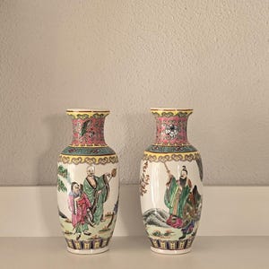 Pode incluir: Dois vasos de porcelana a condizer com cenas pintadas à mão de figuras com trajes tradicionais. O pescoço de cada vaso é decorado com um padrão floral rosa, enquanto o corpo exibe ilustrações coloridas de pessoas num cenário paisagístico.