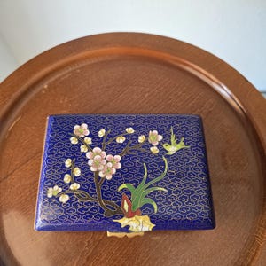 Op de afbeelding: Een rechthoekige cloisonné doos met een diepblauwe achtergrond en een gouden draadpatroon van wolken. Het deksel toont een kleurrijk emaille ontwerp van een bloeiende tak met roze en witte bloesems, groene bladeren en een kleine vogel in vlucht.