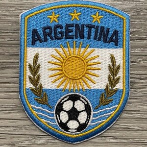 Op de afbeelding: Een schildvormige geborduurde patch met de tekst ARGENTINA in donkerblauw, drie gele sterren, een centrale gele zon, twee gouden lauriertakken en een zwart-witte voetbal, omrand door een dikke gele en blauwe rand.
