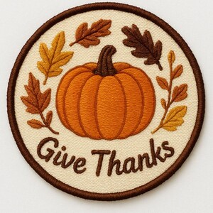 May include: A circular embroidered patch featuring an orange pumpkin in the center surrounded by brown and yellow autumn leaves on a cream background. The text Give Thanks is stitched in brown thread below the pumpkin with a brown border.
