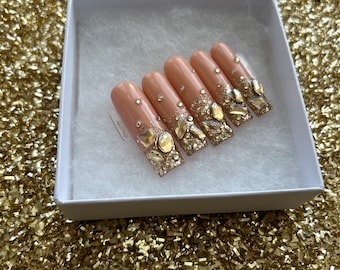 May include: A set of five nude-colored press-on nails with gold glitter and rhinestones.