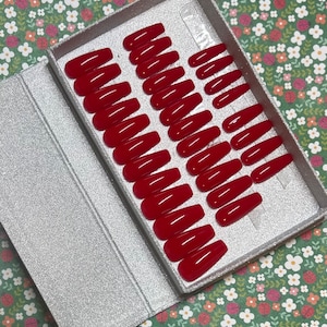 Puede incluir: Un juego de 24 uñas postizas rojas en forma de ataúd en una caja plateada con un inserto de plástico transparente. Las uñas están dispuestas en filas y están listas para ser aplicadas.