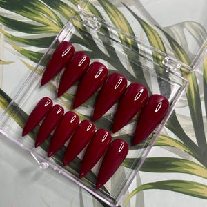 May include: A set of ten burgundy stiletto false nails in a clear plastic box. The nails are arranged in two rows of five.