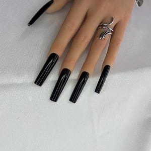 May include: A hand with long, glossy black artificial nails. A silver snake ring wraps around the ring finger. The nails are a solid, rectangular shape and have a high-shine finish. The background is a white, textured fabric.