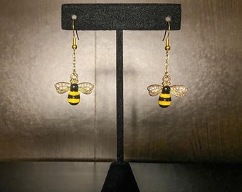 Può includere: Un paio di orecchini a forma di ape dorati con strisce nere e gialle e strass trasparenti. Gli orecchini sono appesi a un espositore per orecchini nero.
