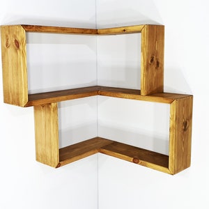 May include: A wooden corner shelf unit with three shelves. The shelves are made of natural wood with a light brown stain. The shelves are in a corner of a room.