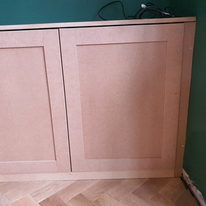 K&ouml;nnte beinhalten: Zwei Holzt&uuml;ren mit hellbrauner Oberfl&auml;che. Die T&uuml;ren sind geschlossen und haben ein schlichtes Design mit einem erhabenen Feld. Die T&uuml;ren sind an einer Wand mit gr&uuml;nem Hintergrund montiert.