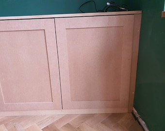 May include: Two wooden cabinet doors with a light brown finish. The doors are closed and have a simple design with a raised panel. The doors are mounted on a wall with a green background.