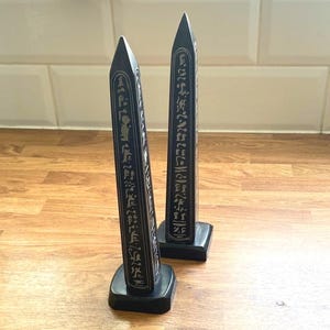 Könnte beinhalten: Zwei hohe, schwarze, vierseitige Obelisk-Skulpturen auf quadratischen Sockeln. Jeder Obelisk weist vertikale Spalten mit eingravierten, hellen, hieroglyphenartigen Symbolen auf allen sichtbaren Seiten auf und läuft oben spitz zu.