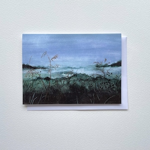 May include: A watercolor greeting card featuring a landscape scene. The artwork depicts a field of tall grass and plants in shades of green and black, with a misty mountain range in the background under a blue sky.