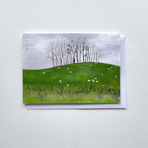 Puede incluir: Una tarjeta de felicitación con una pintura de acuarela de una colina verde con un grupo de árboles desnudos bajo un cielo gris. Pequeños puntos blancos están dispersos por la colina, y hierba alta está en primer plano. La tarjeta está sobre un fondo blanco.