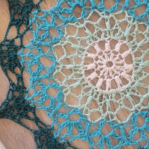 May include: A white and green crocheted dream catcher with a seashell hanging from the bottom. The dream catcher has a circular frame and a woven design with a gradient of colors from dark green to light green to white.