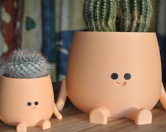 May include: Two orange ceramic planters shaped like people with smiling faces and black eyes. The larger planter has two cacti plants inside. The smaller planter has a single cactus plant inside.