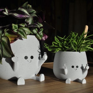 Peut inclure: Deux pots de fleurs en céramique blanche en forme de personnes avec les bras levés en signe de paix. Les pots ont des yeux noirs et une bouche souriante. Le plus grand pot contient une plante verte et violette, tandis que le plus petit pot contient une plante succulente verte.