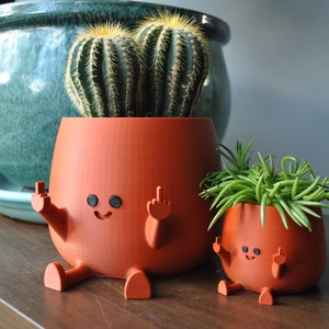 Puede incluir: Dos macetas de cerámica naranja con forma de personas con brazos y piernas, haciendo el dedo medio. La maceta más grande tiene una planta de cactus dentro y la maceta más pequeña tiene una planta suculenta verde dentro.