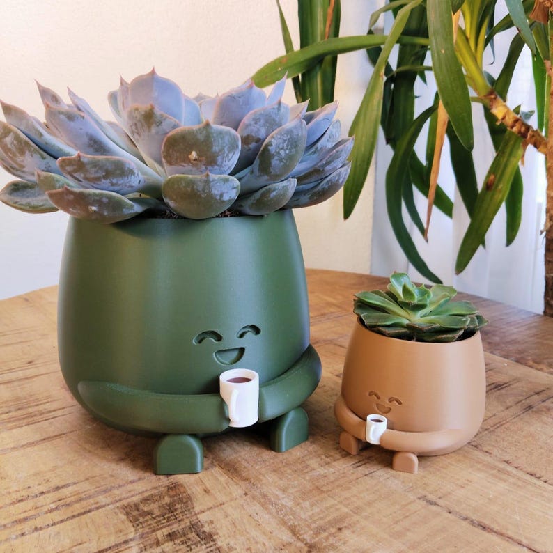 May include: Two novelty plant pots, one olive green and one tan, each with a smiling face and arms holding a small white mug. The larger pot contains a blue-green succulent. The pots are designed to look like they are hugging the mugs.