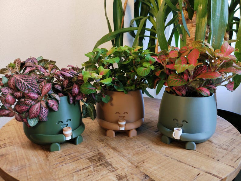 May include: Three decorative plant pots in green, brown, and olive green, each holding a small plant with colourful foliage. The pots have a smiling face design and are holding a small coffee cup. The pots are sitting on a wooden table.