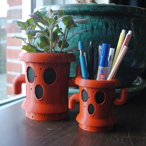 May include: Two orange ceramic planters shaped like cartoon characters with black eyes and mouths. The planter on the left has a plant with green and red leaves. The planter on the right has a variety of pens and pencils inside.