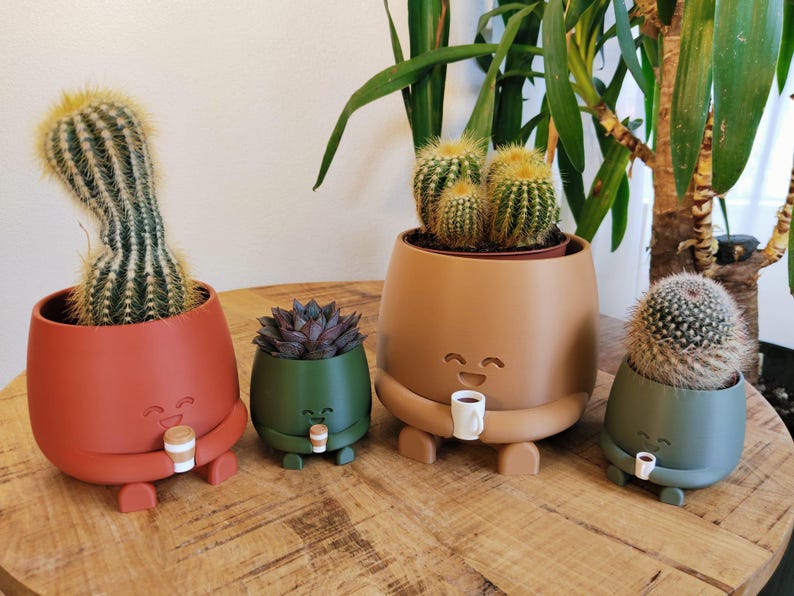 May include: Four quirky cactus pots in red, green, and tan, each with a smiling face and holding a small coffee cup. The pots are on a wooden surface, with various cacti and succulents inside.