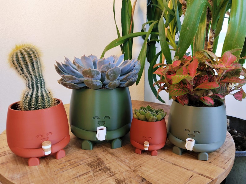 May include: Four quirky plant pots in red and green, each with a smiling face and holding a coffee cup. The pots contain a cactus, succulents, and foliage, set on a wooden surface.