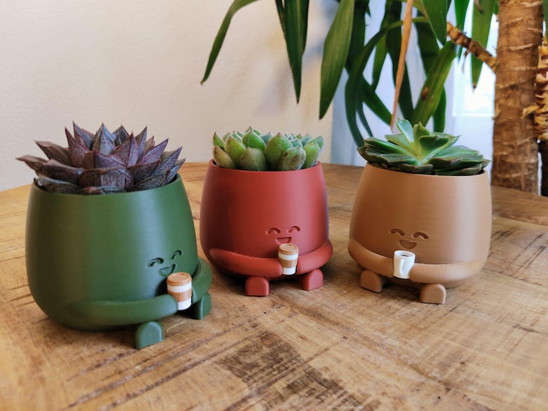 May include: Three colourful succulent planters in green, red, and brown, each with a smiling face and holding a small coffee cup. The planters are on a wooden surface and contain different types of succulents.